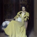 Lady sat in chair looking seductively in armchair with two small fluffy dogs. Lady is wearing long satin yellow dress.