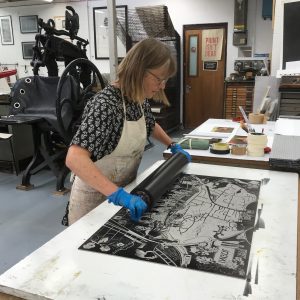 Image shows artist in a studio rolling over a lino print of the Ingol tea towel design.