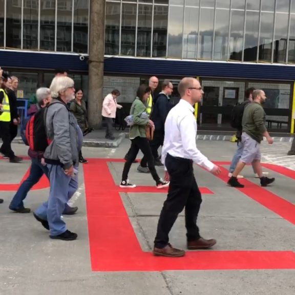 Image shows people walking over a large red sign that says 'People' painted on Preston bus station ground.