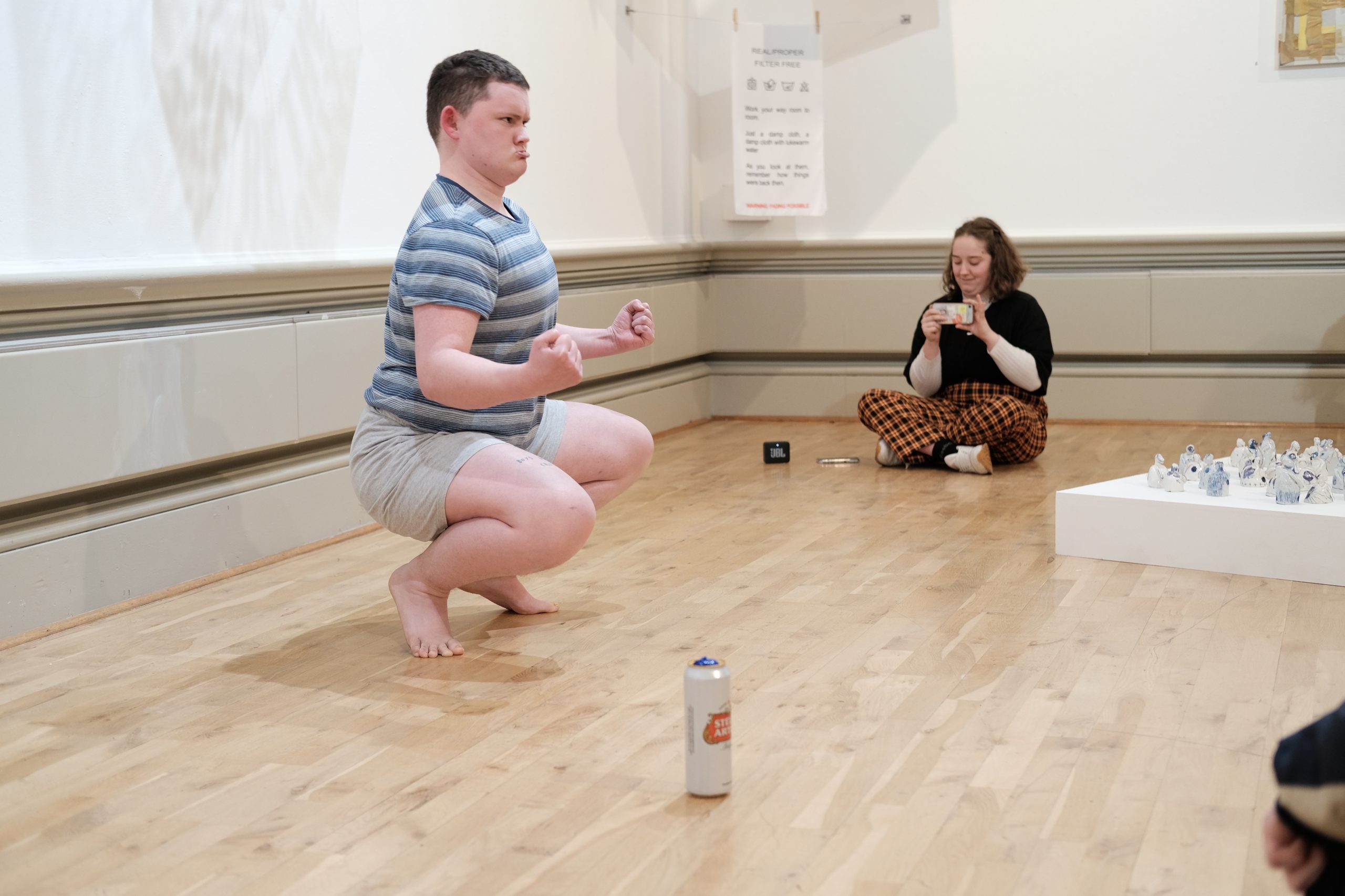 Image of Theo Seddon squating on the floor in the exhibition gallery, clenching his fists and performing part of his piece titled 'Dawn of Man'