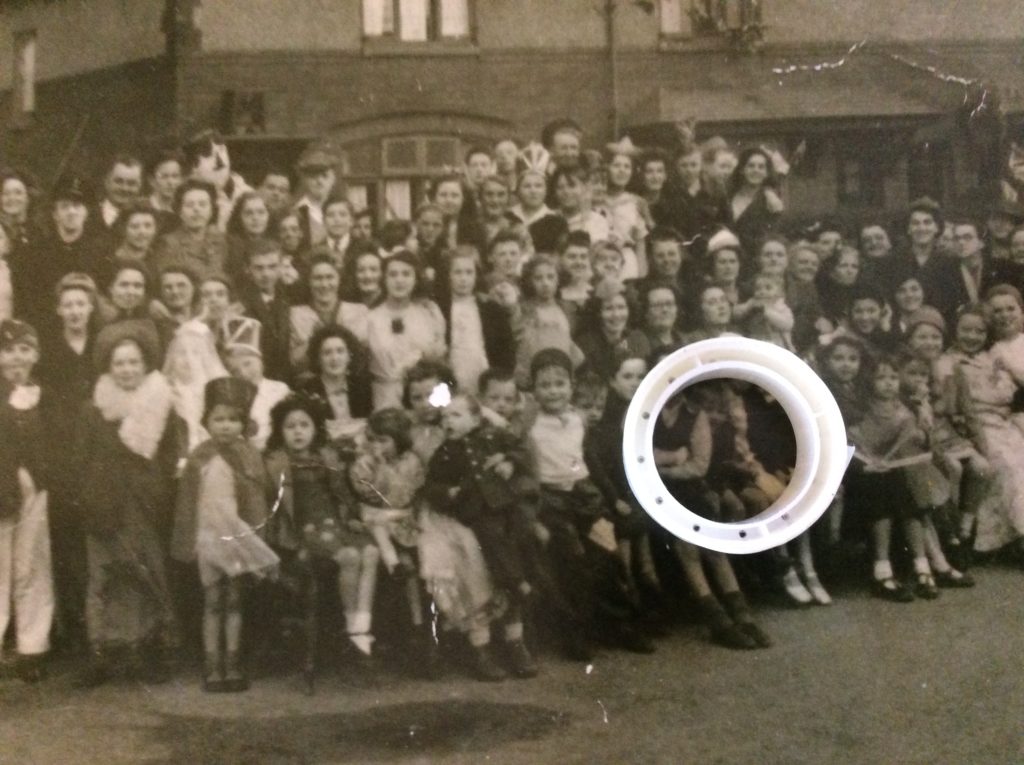 Old photo from the 40s. Lots of children and families stood outside smiling to camera. 
