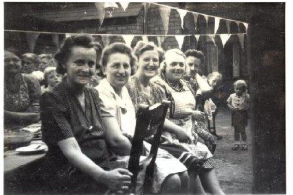 5 ladies at a streeth party with people behind them and bunting decoration overhead. There are 2 children in the background looking towards the camera. The ladies are smiling. 