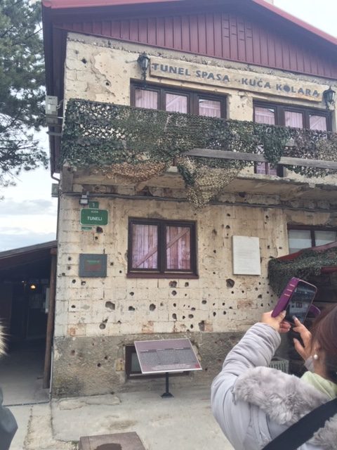 Photograph of a building with bullet holes through its bricks. The words 'Tunel Spasa Kuca Kolara' are painted at the top of the building. 