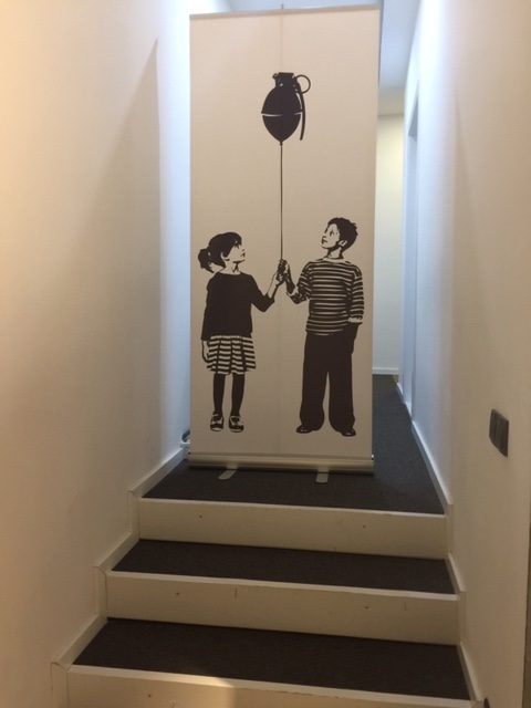 Photo of two children holding a balloon string together. There is a bomb attached to the top of the ballon string. Both children are looking up at the bomb floating above them. This illustration has been printed onto a roll up banner and photographed in this imaged.