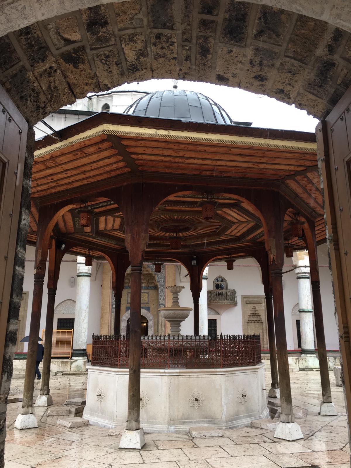 Photo of large structure with a domed roof. The supporting beams of the structure are made of wood with a fountain in the middle. 