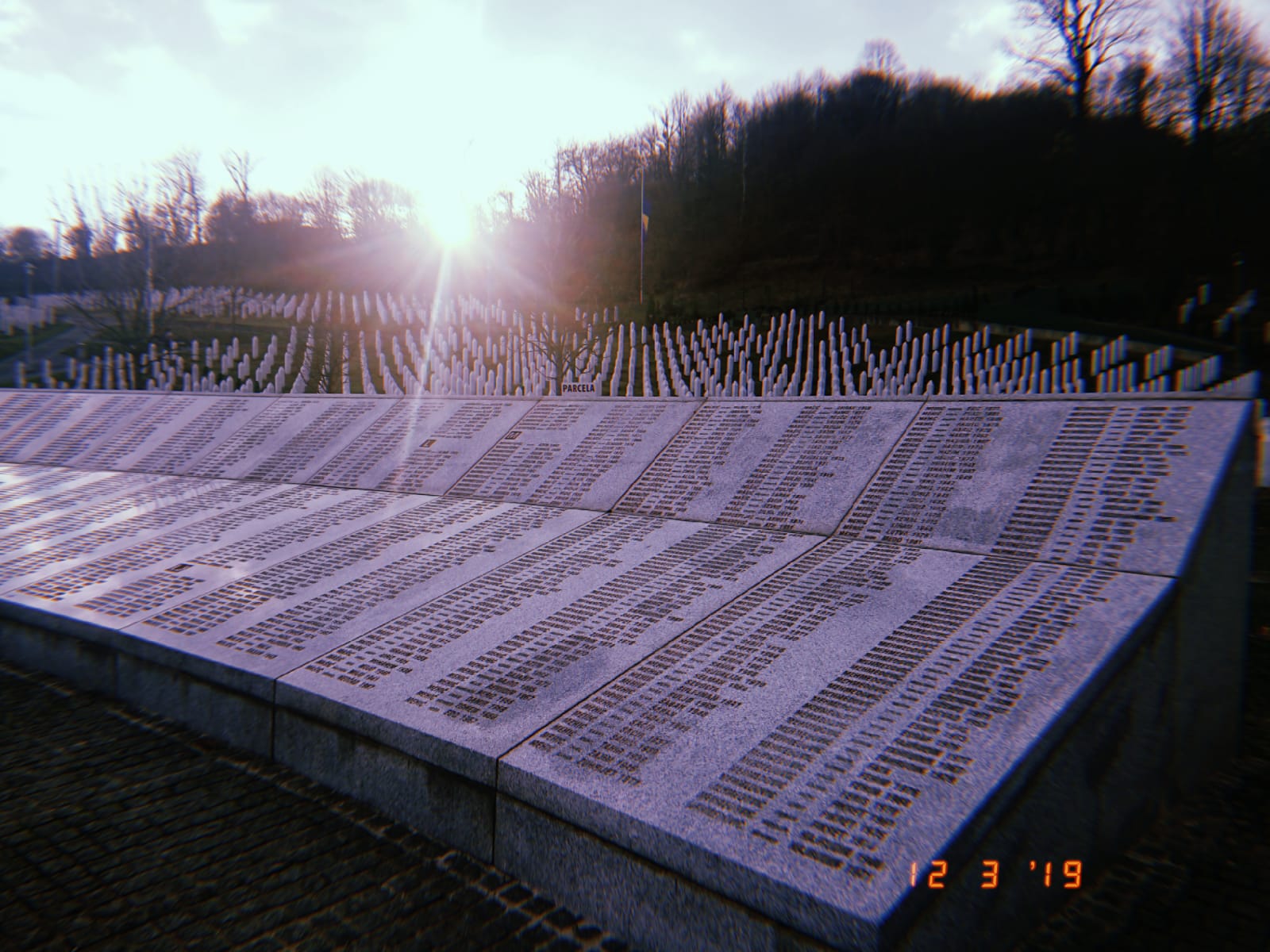 A long list of names of those who died in the Srebrenica Massacre listed on a stone structure. Behind the stone structure is a field with pointed tombstones spreading into the distance. 