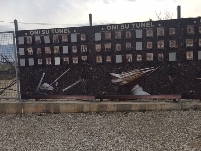 A wall of photos of men with the tools used to build the tunnel laid underneath the photos. The words 'Oni Su Tunel' are inscribed on the wall. 