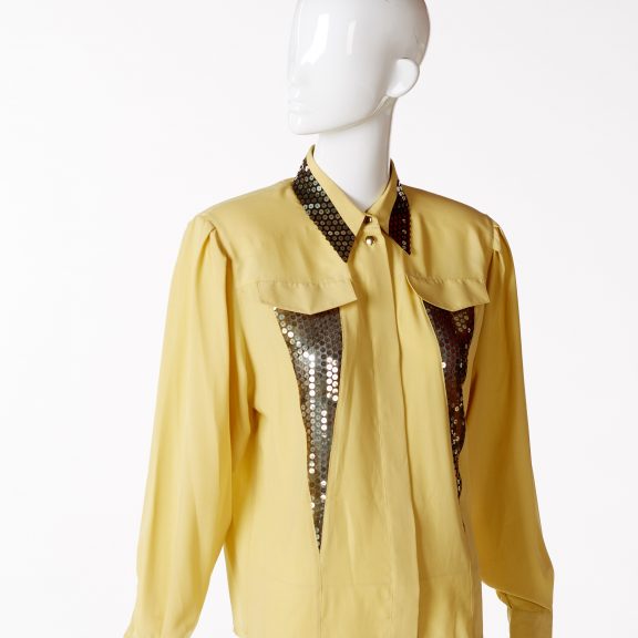 Mannequin wearing mustard yellow evening blouse with long sleeves and a collar. Blouse has appliqued panels of black net and gold sequins - two long triangular panels under the front false pocket flaps on the chest and around edge of collar; fastening down the front with eight buttons.