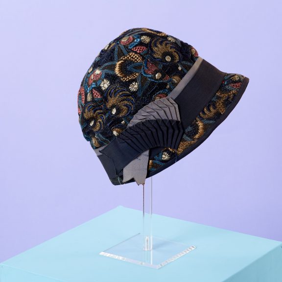 Image shows a cloche hat, embroidered with an all over floral pattern in pink, blue, navy blue, gold and cream, trimmed with a navy blue and pale blue gros-grain ribbon. Hat is shown on a clear perspex stand against a blue background on a blue stand.