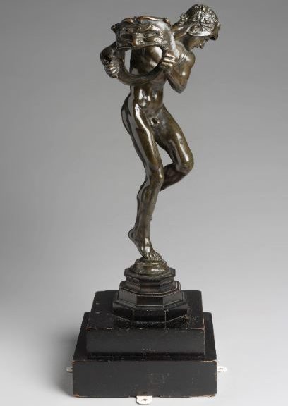 Image shows a bronze sculpture of a naked figure with their face turned away. The figure is holding a large theatrical mask. Sculpture is on a small black plinth base.