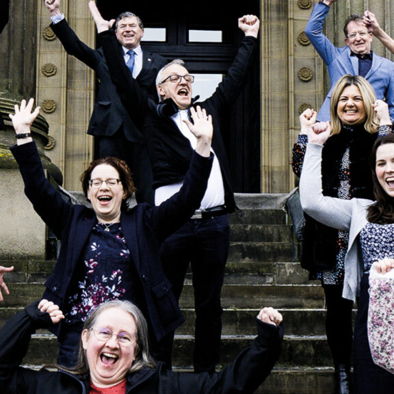 Image fo Harris staff raising their hands in the air pictured outside the front of the Harris