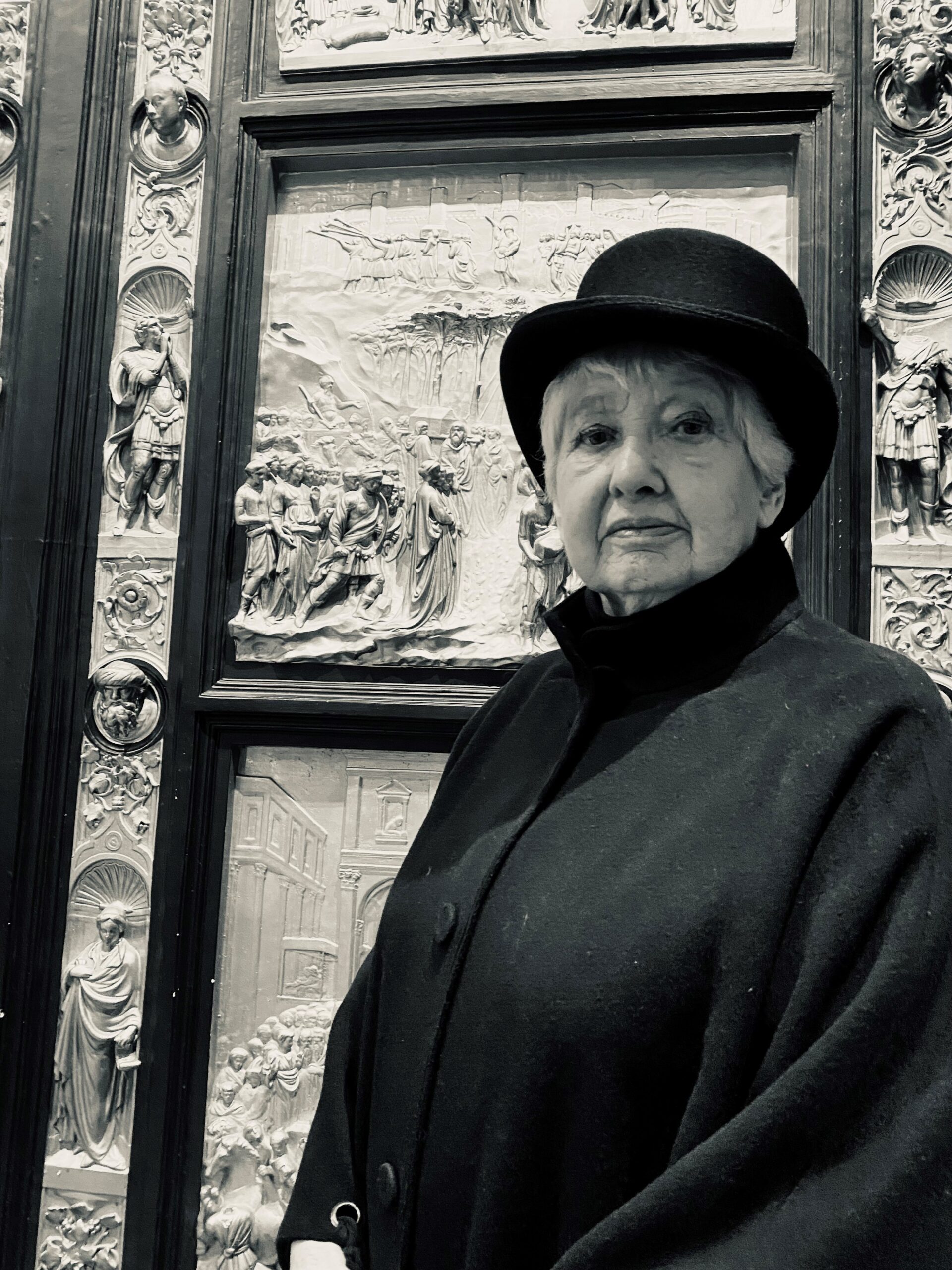 A black and white photograph of a lady wearing black bowler hat and black coat stood in front of an artwork at the Harris