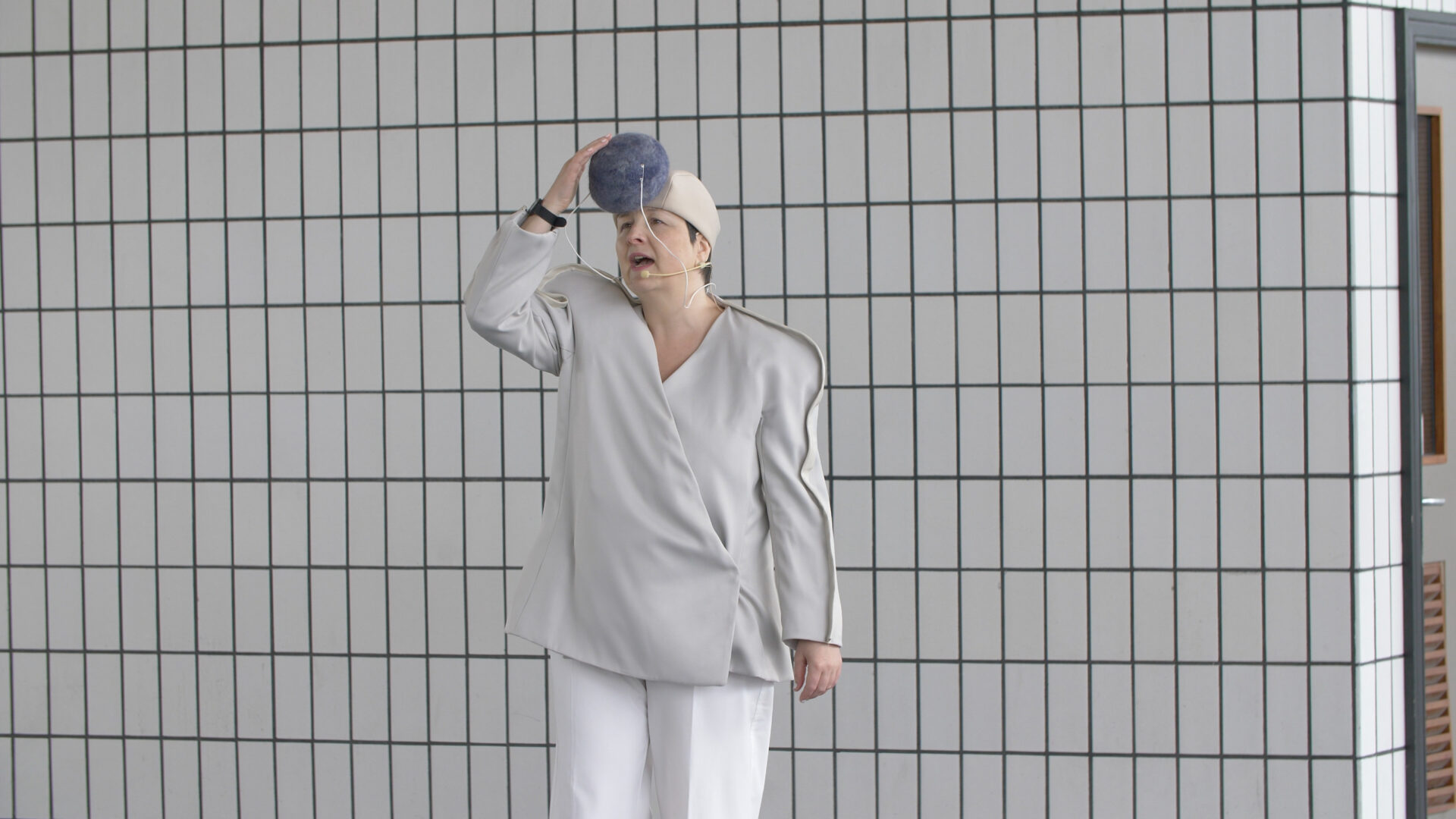 Image taken from a performance at the Bus Station, the artist is dressed in a white sound suit touching a large grey pom pom on their hat.
