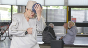 Image taken from a performance at the Bus Station, the artist is dressed in a white sound suit touching a large grey pom pom on their hat.