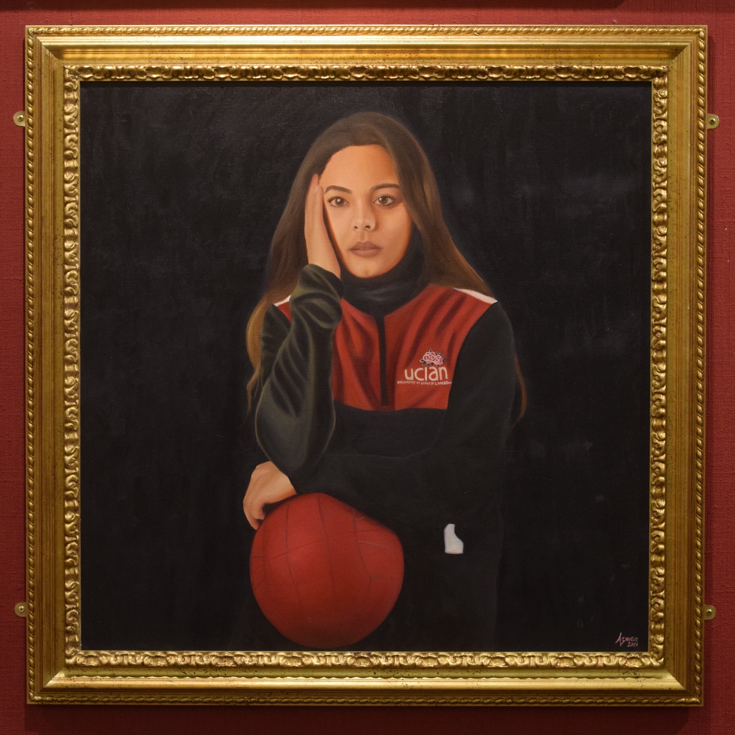 Image of a painting framed in gold of a person leaning on a ball wearing a UCLAN branded jacket.