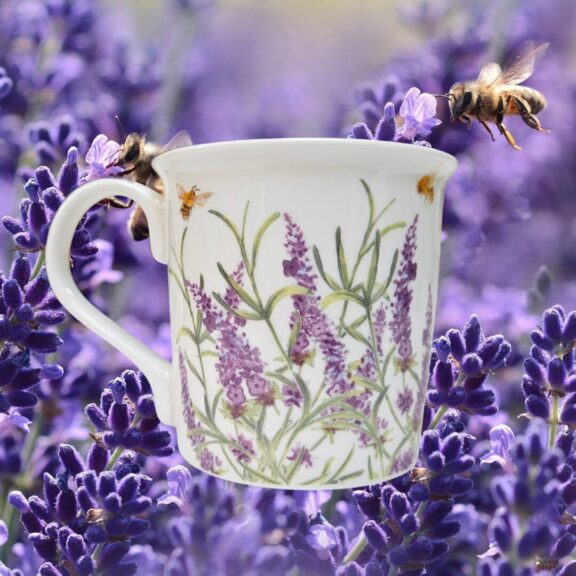 A white ceramic mug decorated with lavender and bees, with lavender in the background.