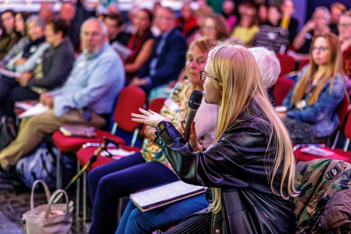 A person speaks into a microphone in front of a group of people sitting down.