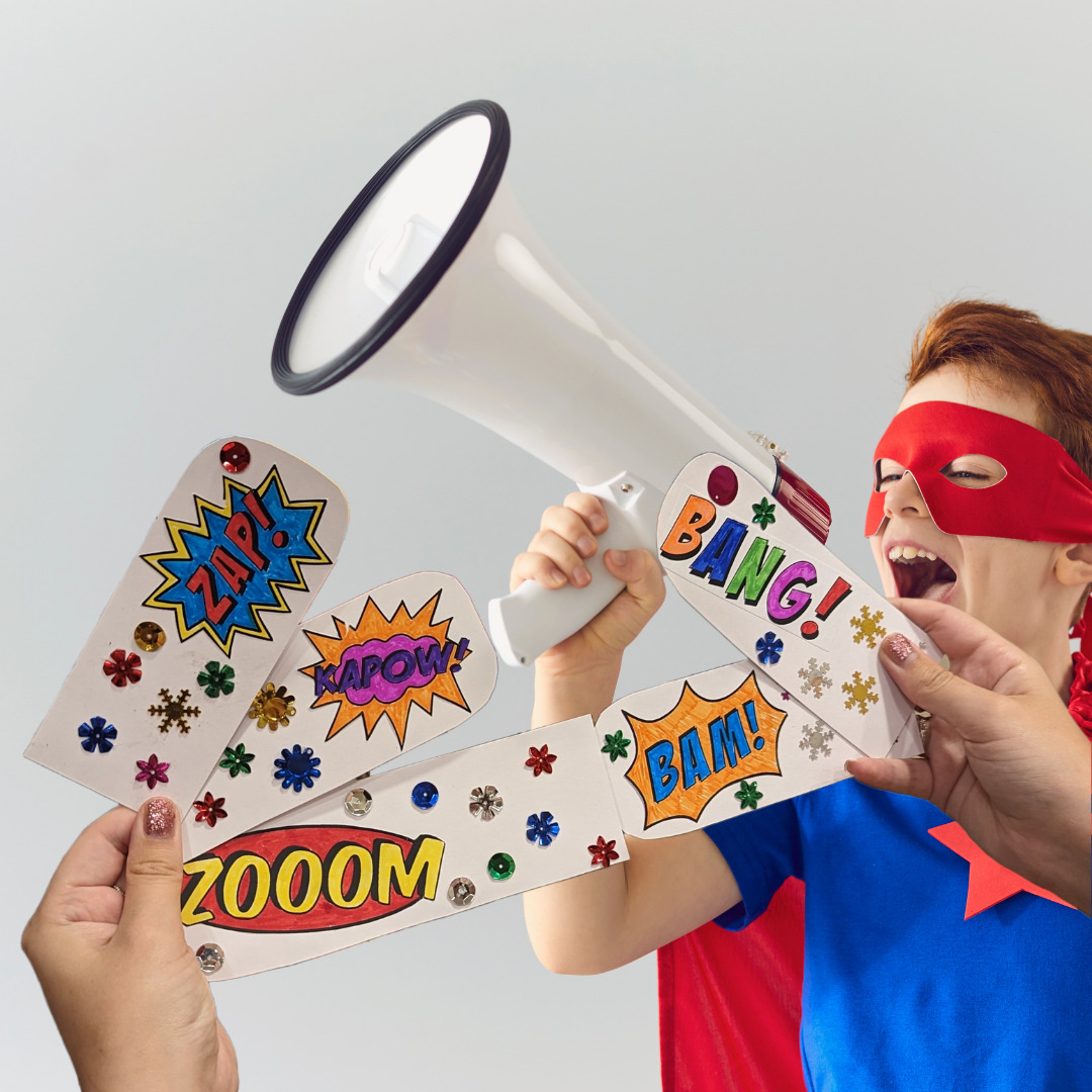 A child dressed in a red and blue superhero costume shouts into a megaphone. Two hands hold up colourful pieces of card.