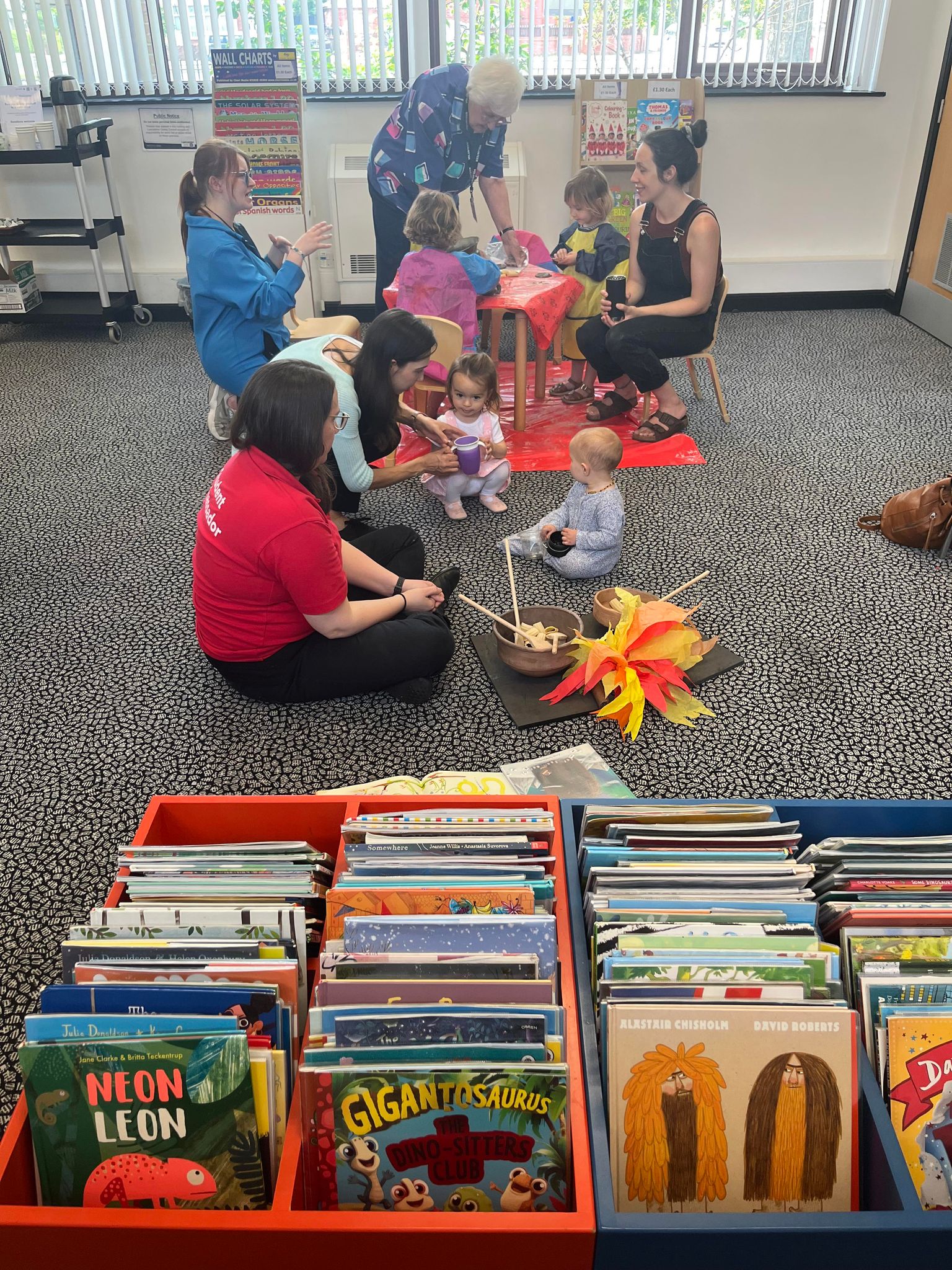 A group of volunteers and children sit and stand around a red table in a library.
