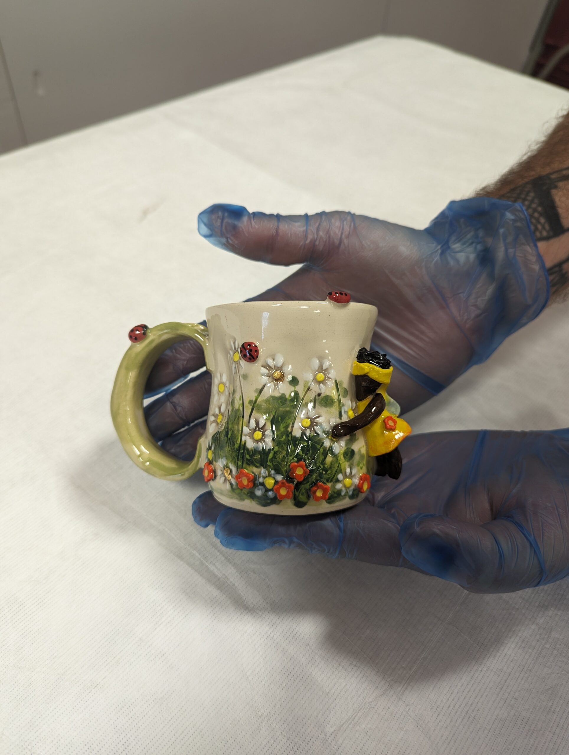 A pair of hands in blue gloves hold a small cream and green ceramic mug decorated with flowers and a fairy.