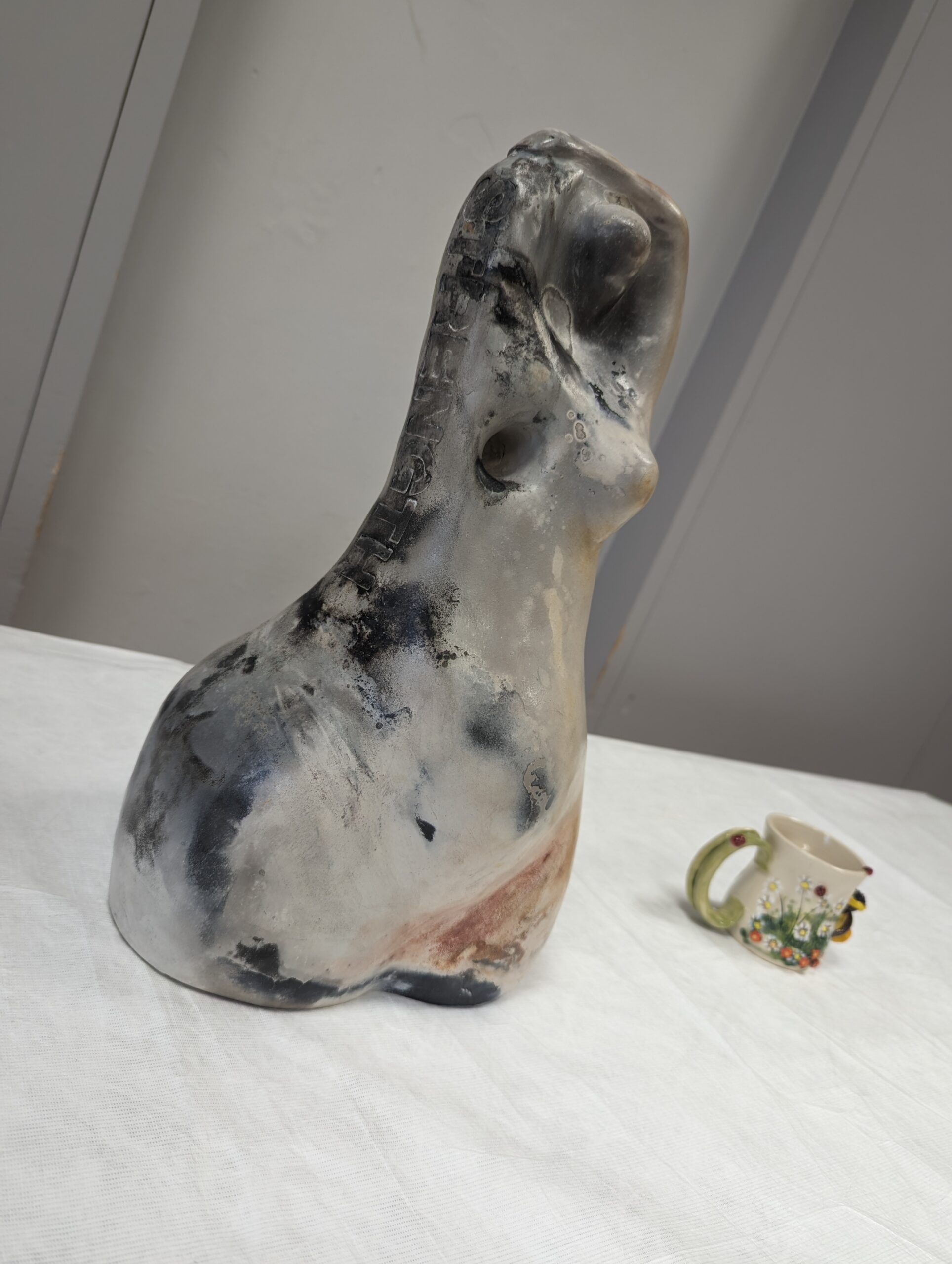 A silver and grey abstract statue of a woman holding her arms above her head, there is a hole where the right breast should be. Next to it is a small cream and green ceramic mug decorated with flowers and a fairy.