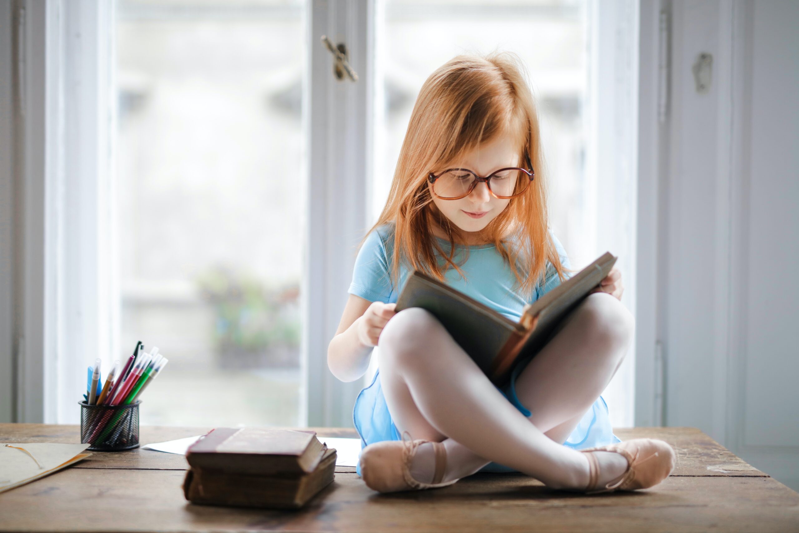 A child with long hair and glasses sits on top of a table reading a book.