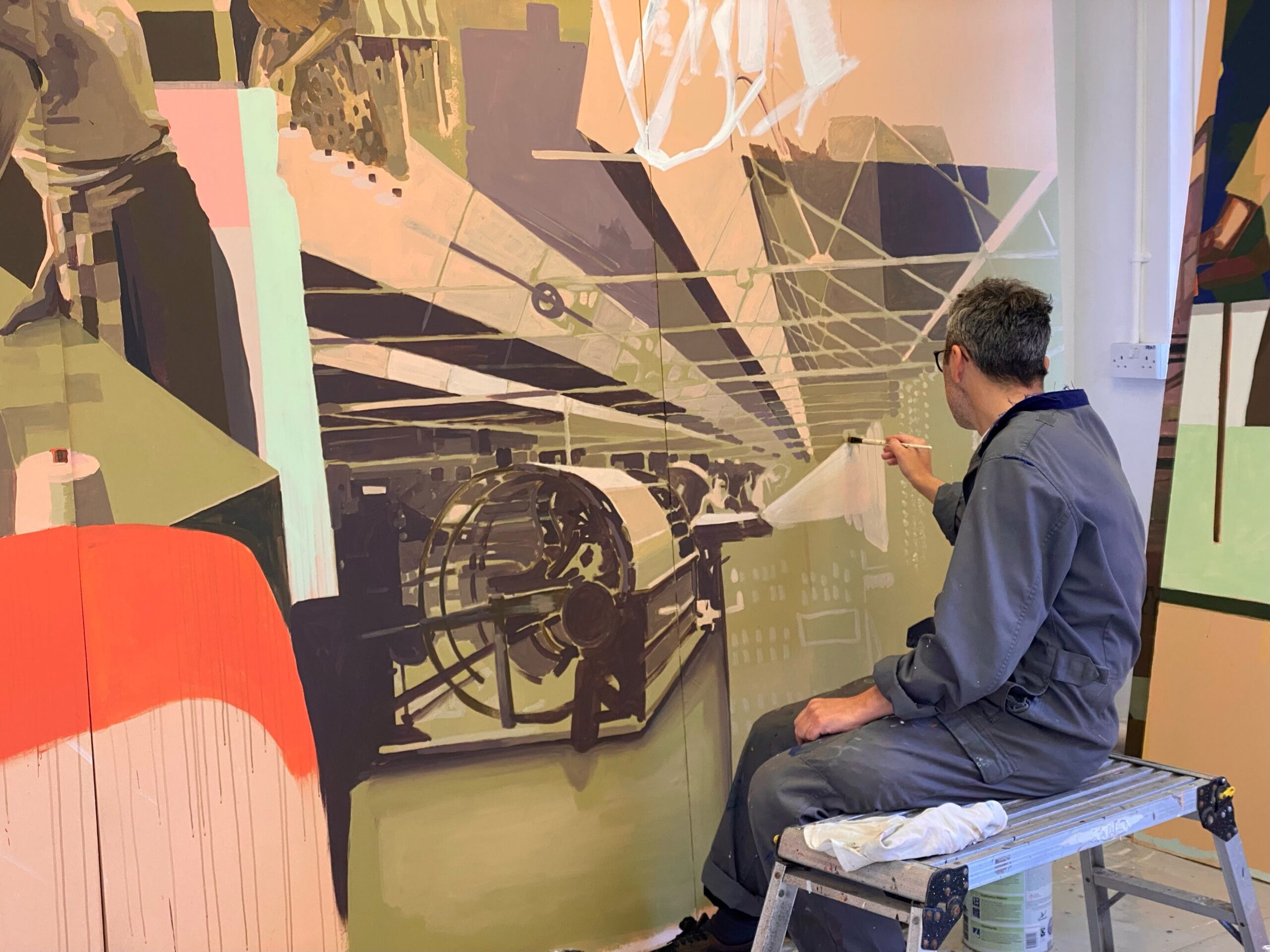 Gavin, dressed in grey overalls, sits on a metal stand as he paints a mural.