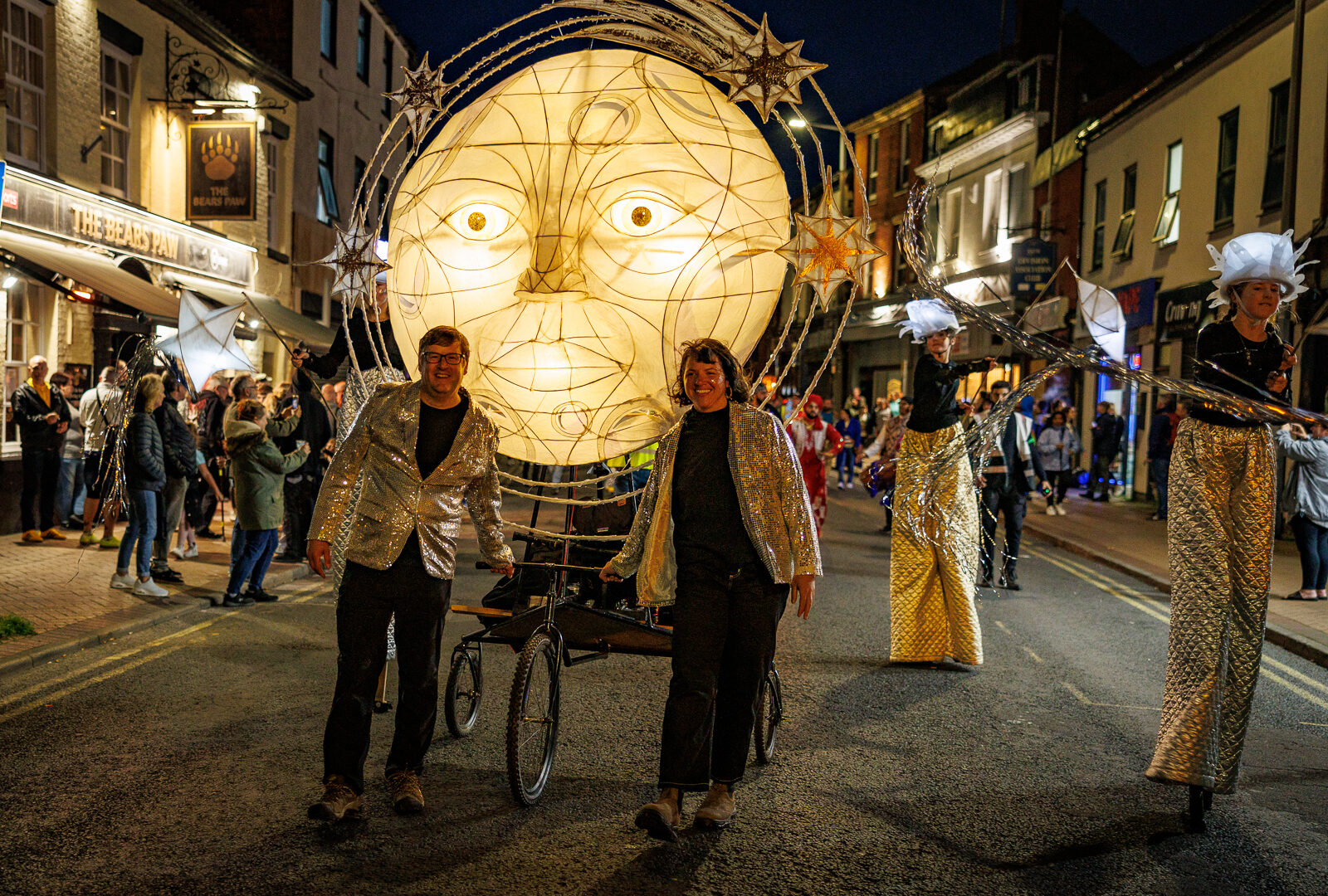 Image of a giant moon light up puppet being pulled by two people wearing sequined jackets