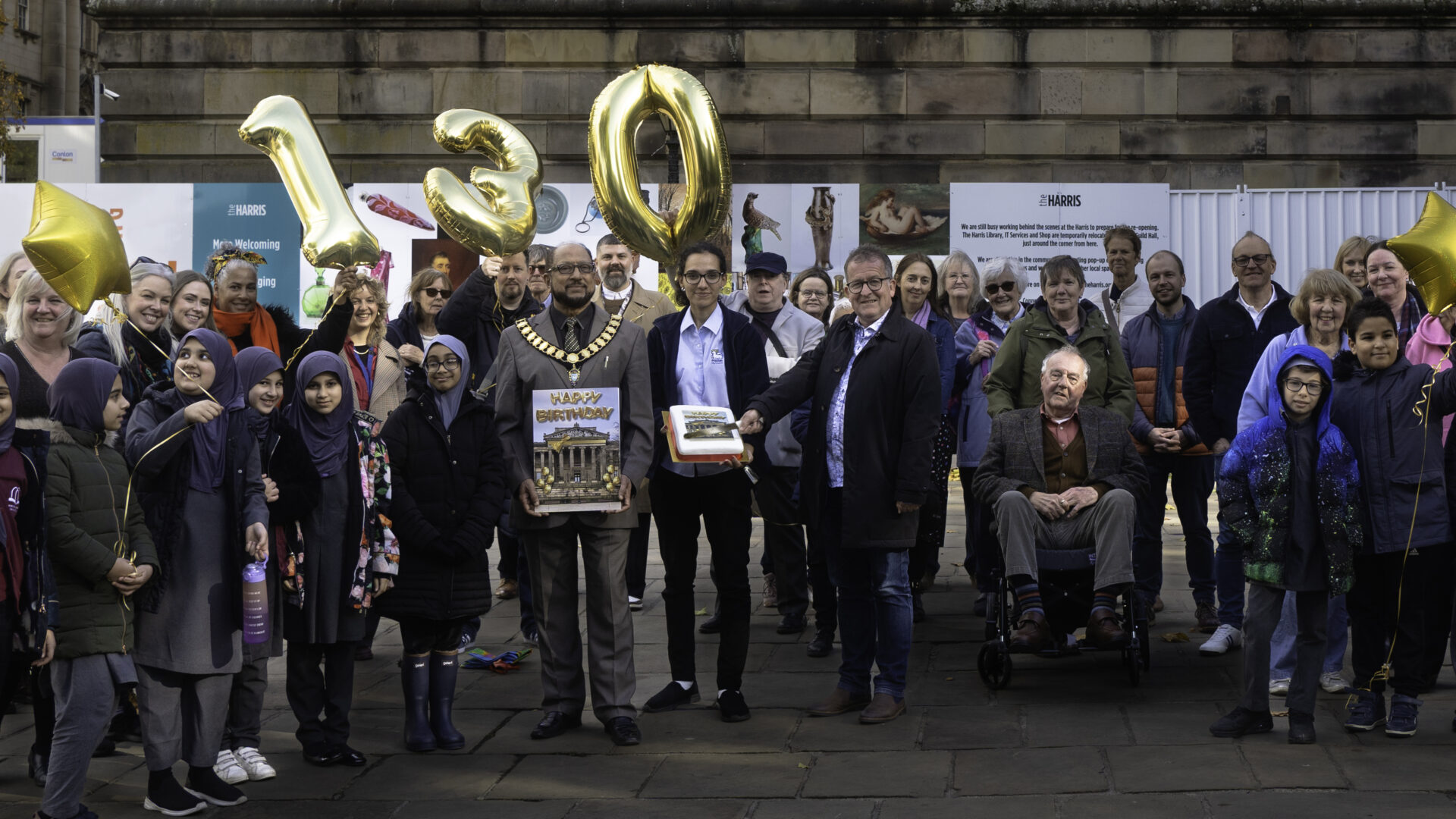 Image of a group of people holding a cake ad 130 balloons.