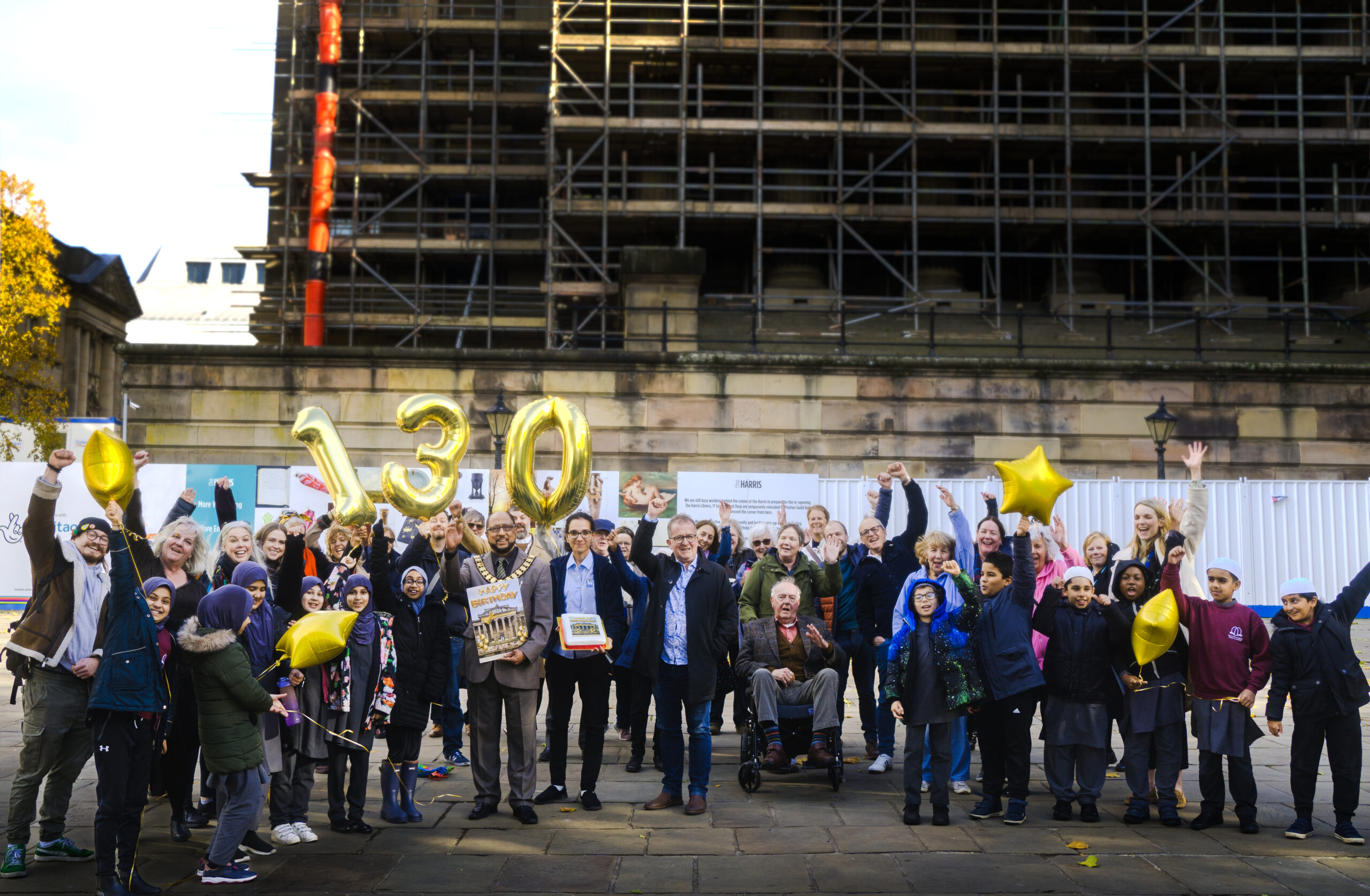 Image of a group of people holding a cake ad 130 balloons.