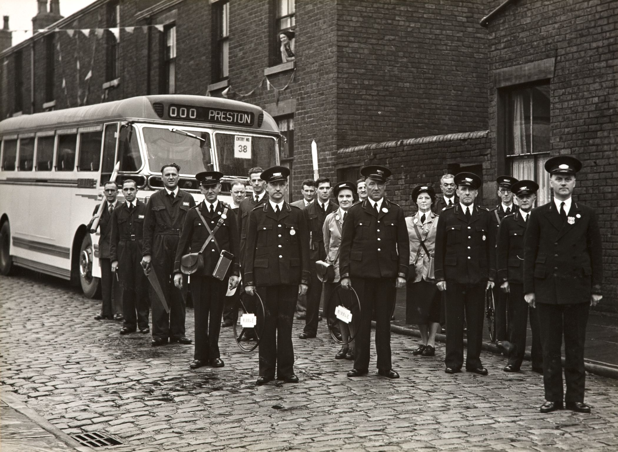 A black and white photograph of bus drivers stood in front of a bus on a cobbled street.