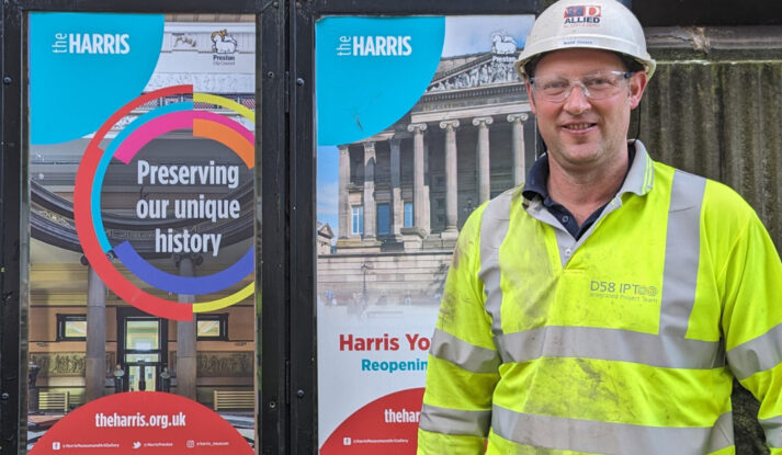 A person in a high vis jacket and a hard hat stands next to two colourful posters outside the Harris