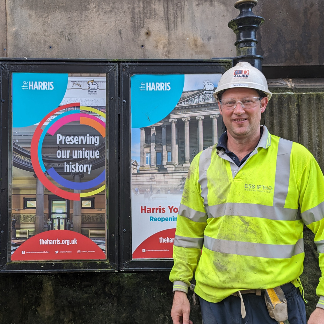 A person in a high vis jacket and a hard hat stands next to two colourful posters outside the Harris