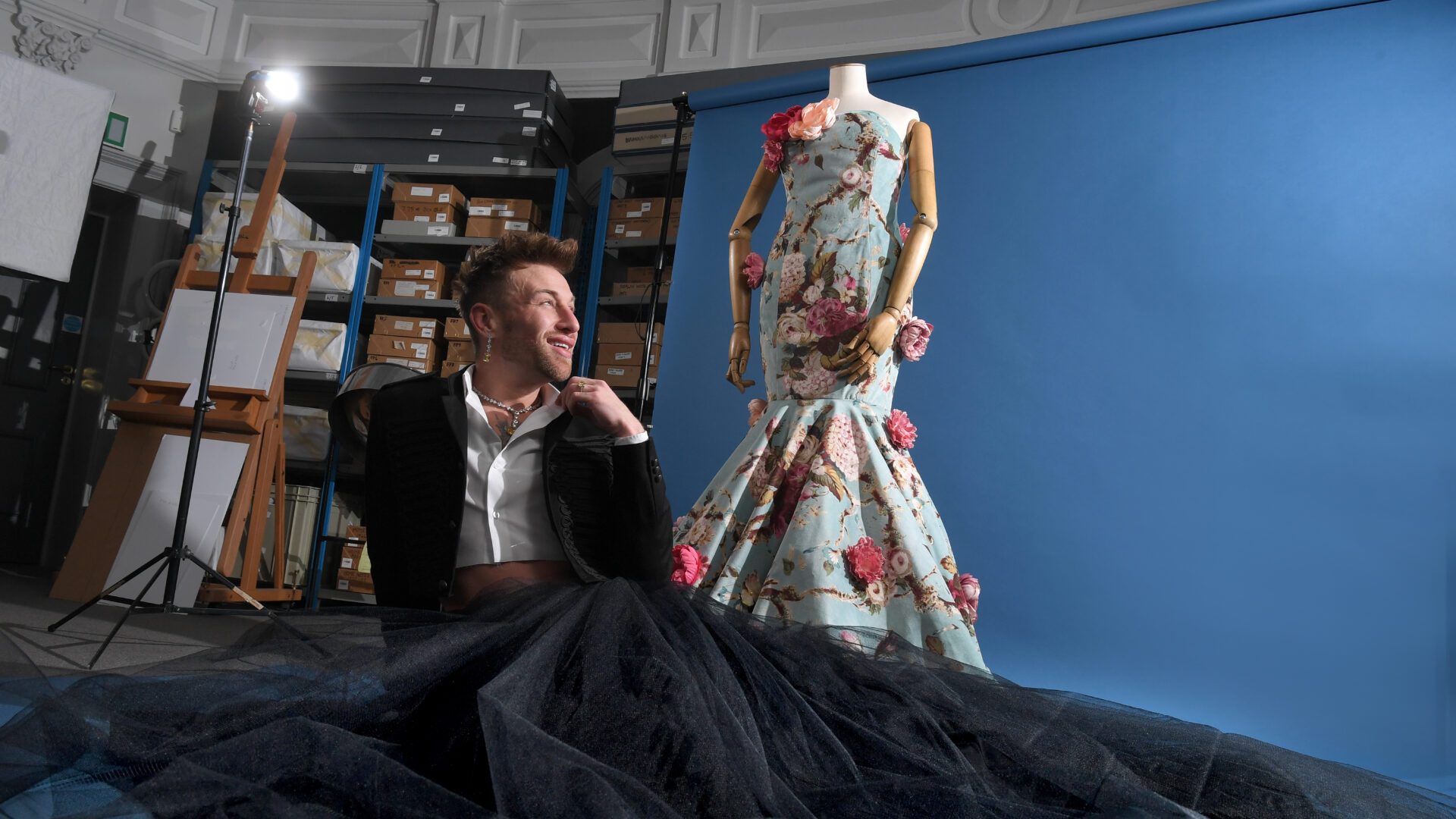 A person wearing a cropped jacket and long skirt sits in front of a rose dress with a blue backdrop.