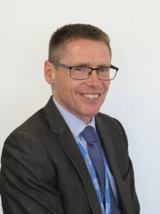 Headshot image of Simon Nixon, a man who wears glasses and a black suit with a blue tie and ‘Preston College’ lanyard around his neck.