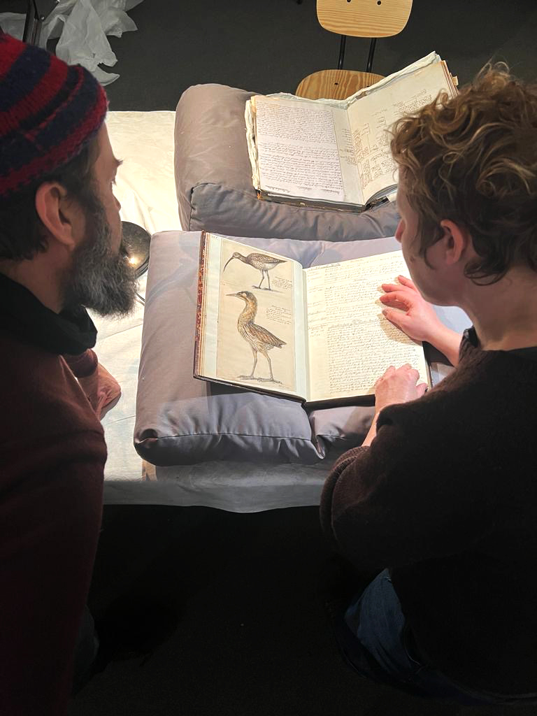 Two people lean over a book displaying illustrations of birds.