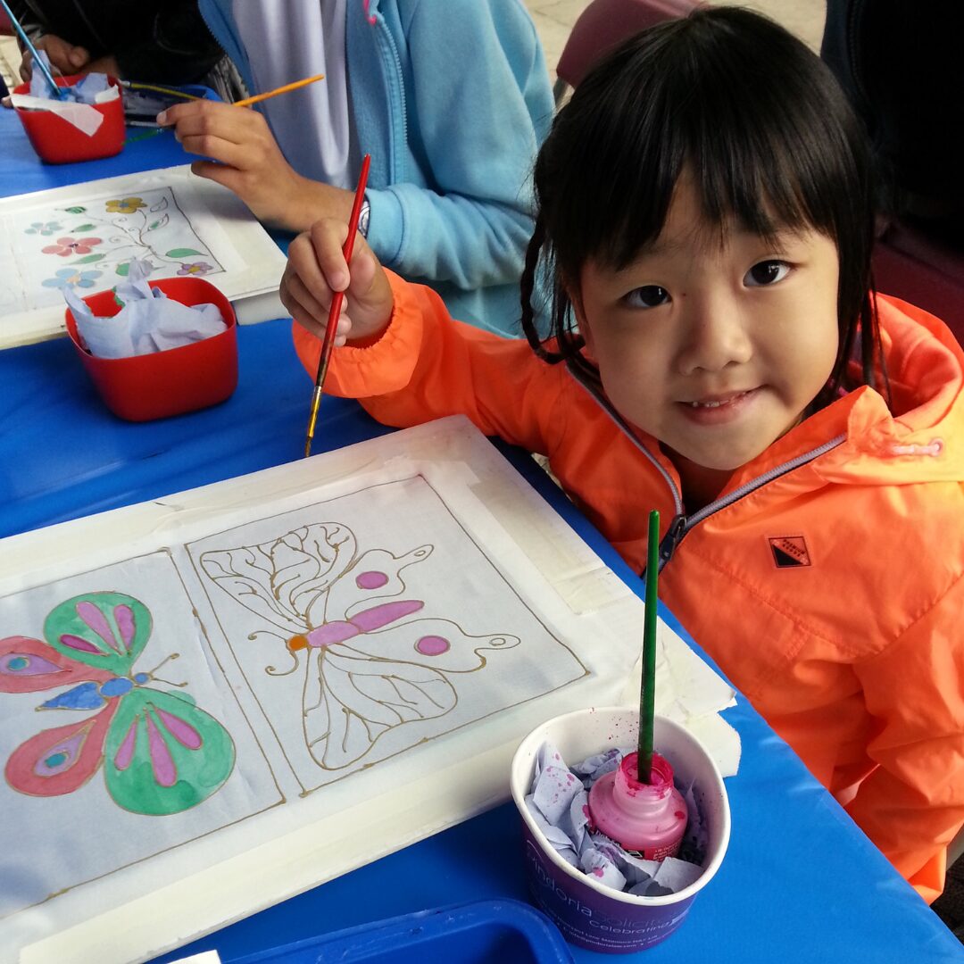 A child looks at the camera whilst painting a butterfly on a piece of silk
