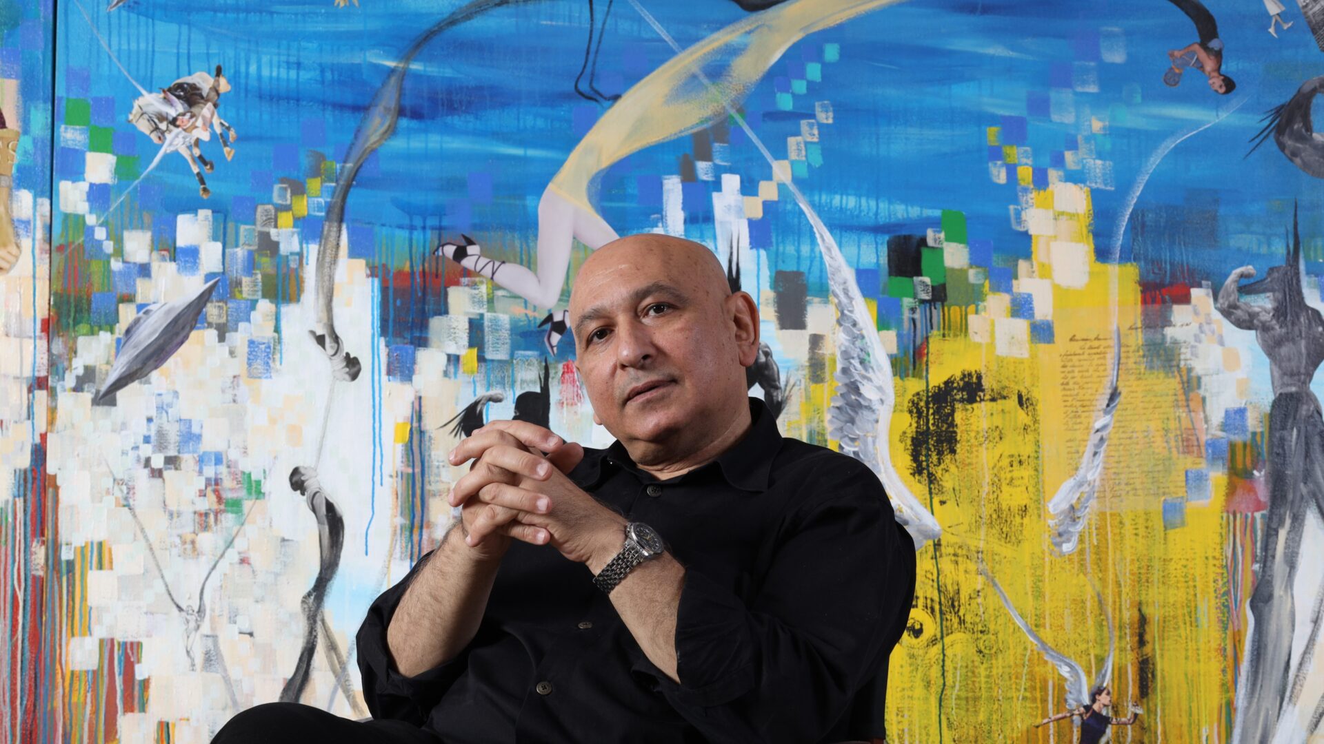 Artist headshot of a person with their hands clasped together in front of a colourful artwork
