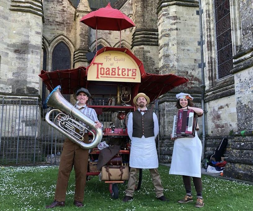 Three people with instruments stood in front of a 'Guild of Toasters' set.